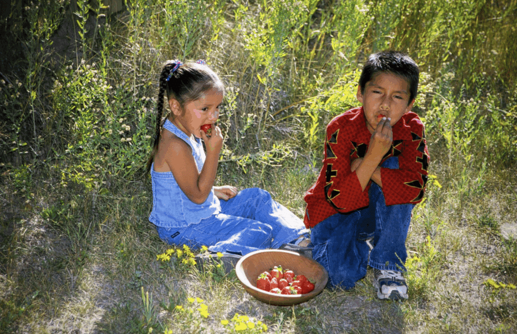 Two Indigenous children sitting in grass eating from a bowl of strawberries