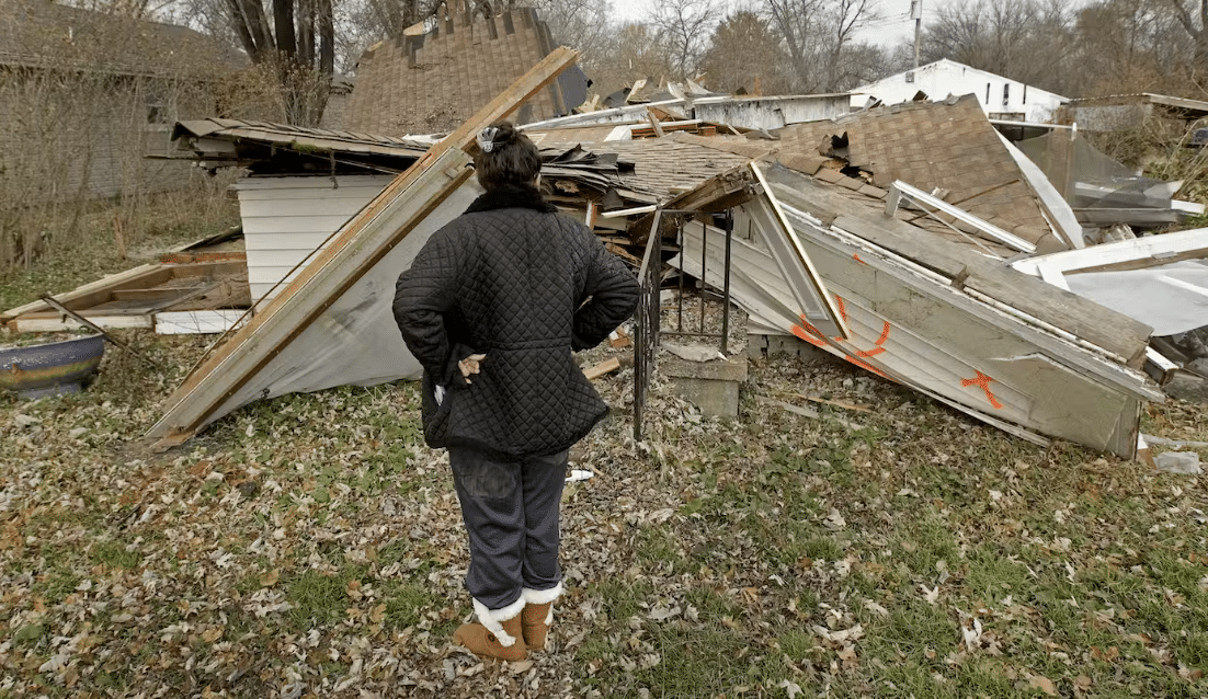 Person stands with hand on hips looking at a home destroyed by a natural disaster