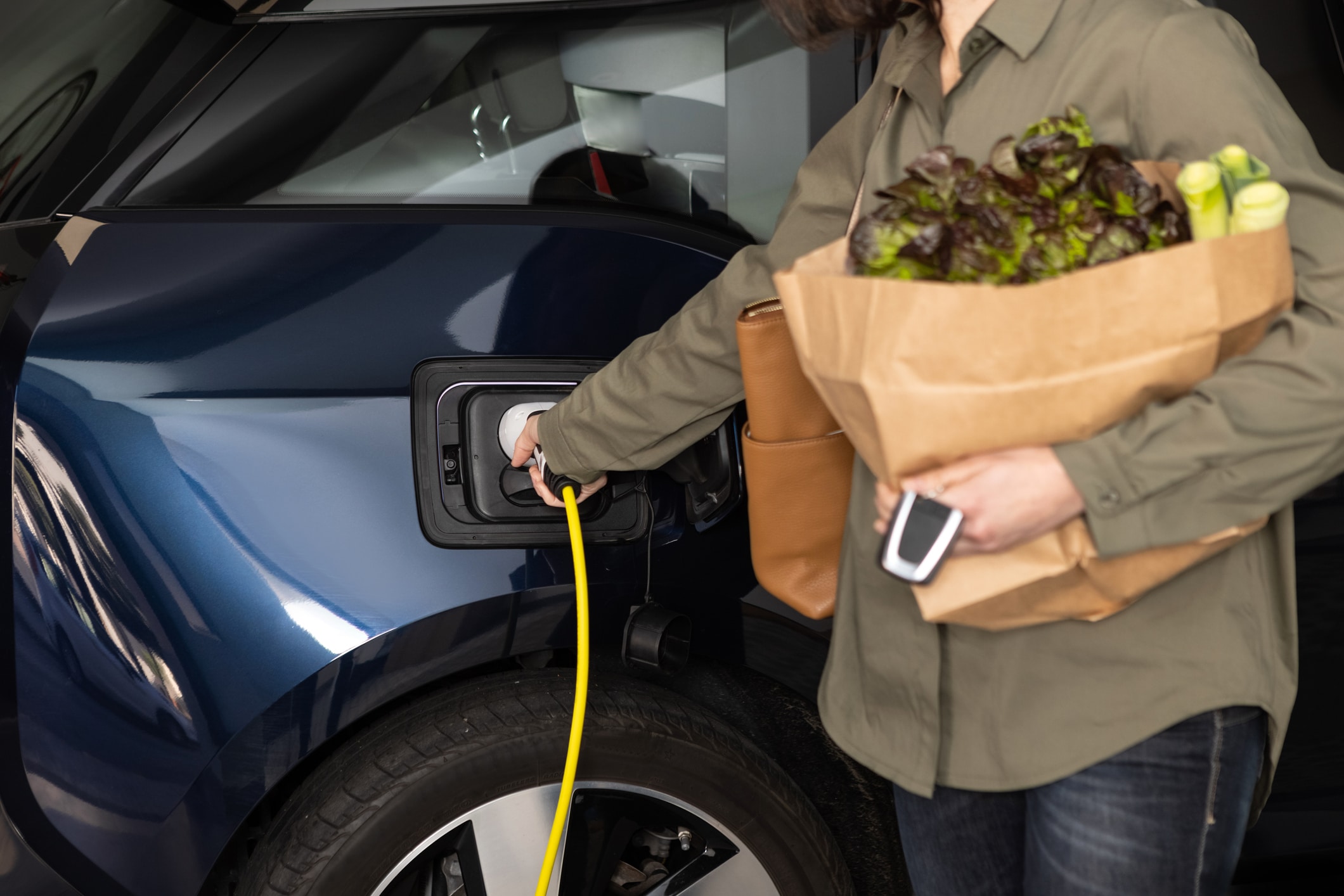 Woman plugs a charger to electric car socket at home after shopping groceries.