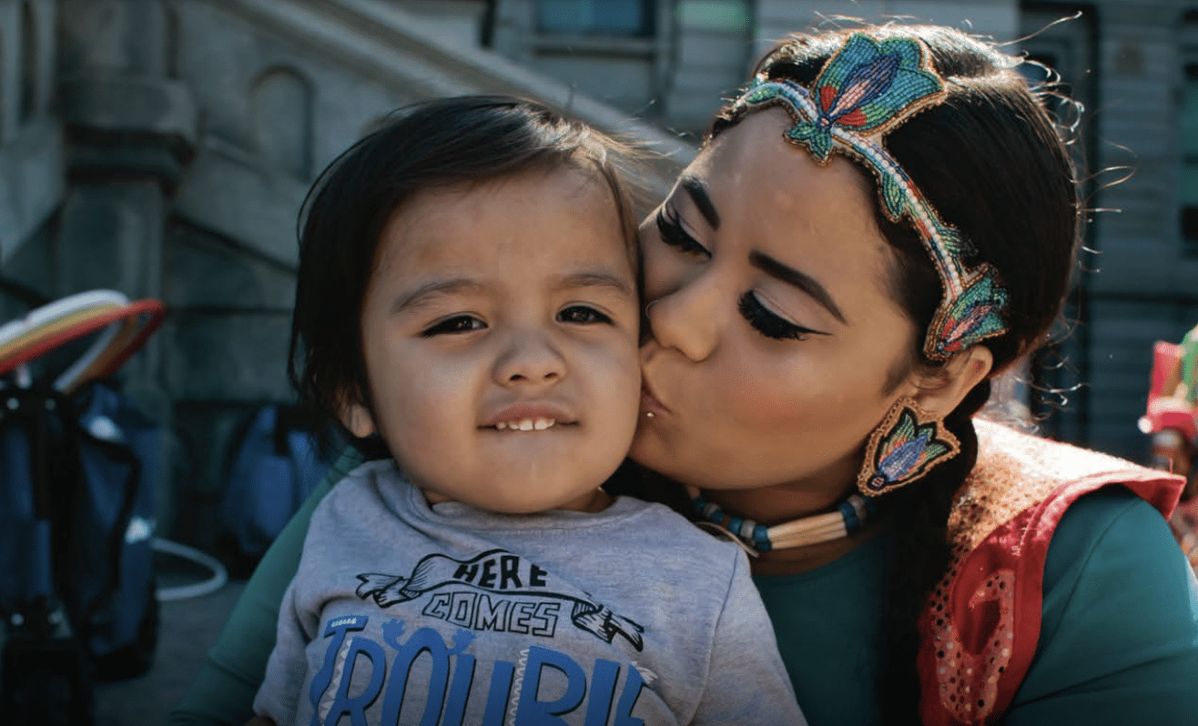 Mother in traditional Native American garb kisses her toddler's cheek