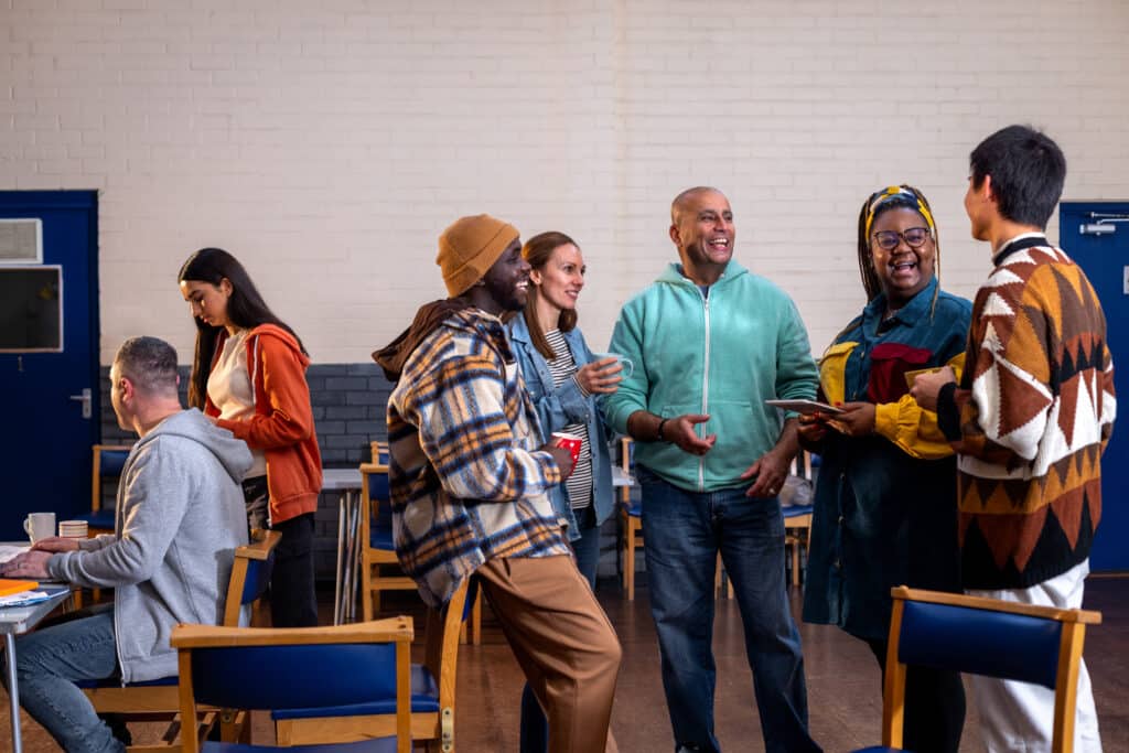 A group of adults standing in a circle and talking to each other about their mental well-being. There is a staff member sitting to the left of them in the frame sitting next to a table. 