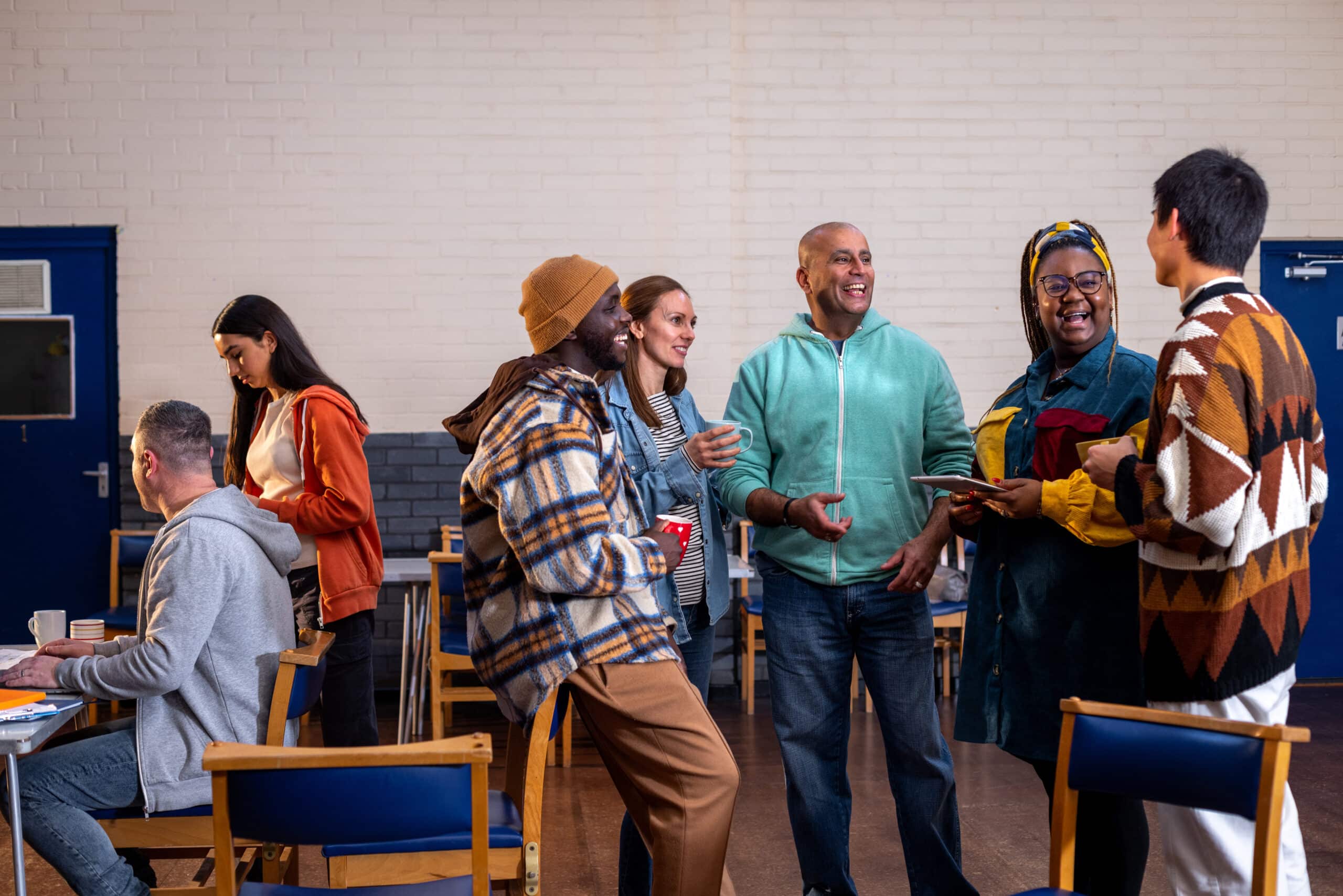 A group of adults engages in animated conversation and networking in a bright community room, while others focus on materials at nearby tables.