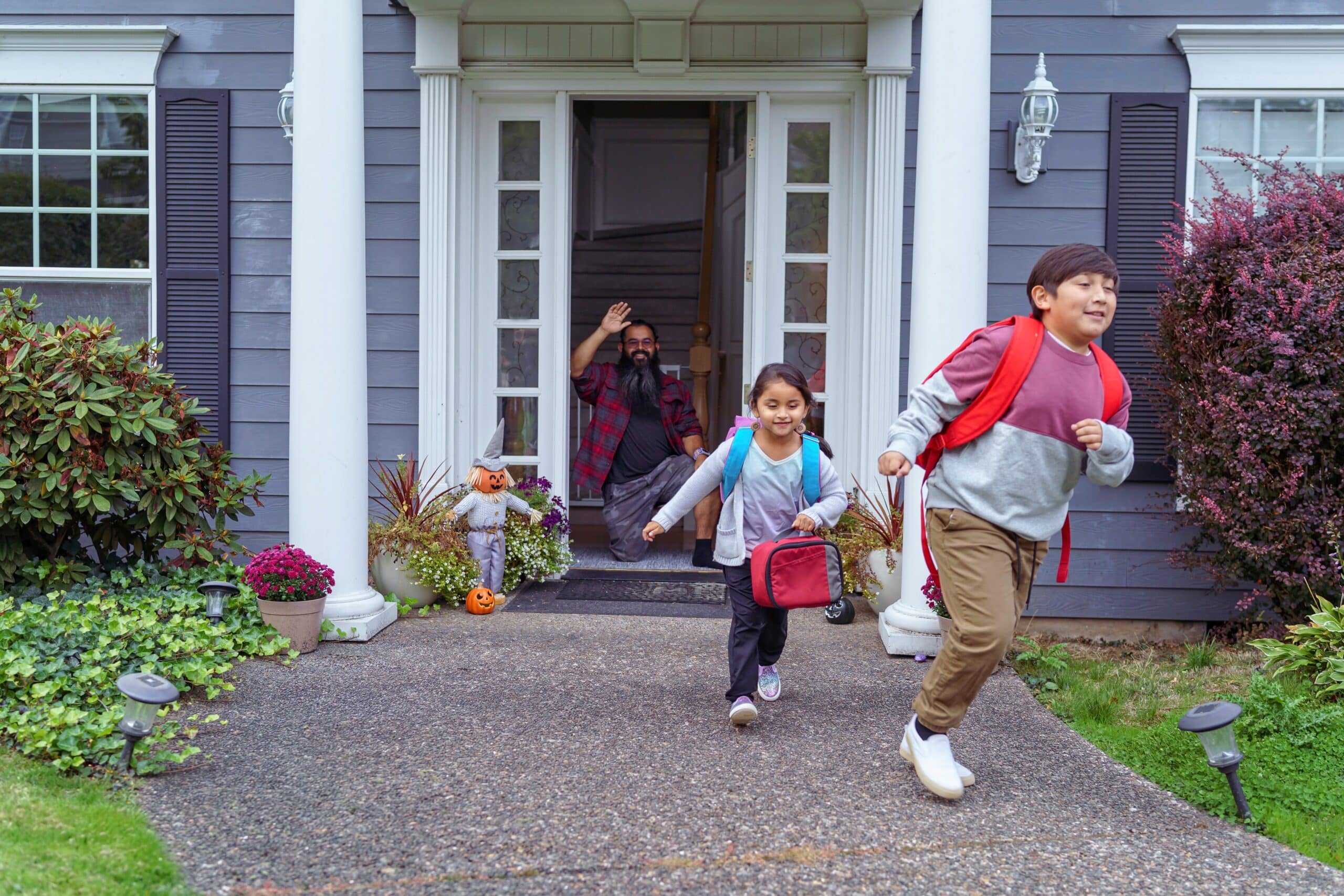 Elementary age Native American brother and sister run to meet the school bus as their dad waves goodbye from the front door of their home.