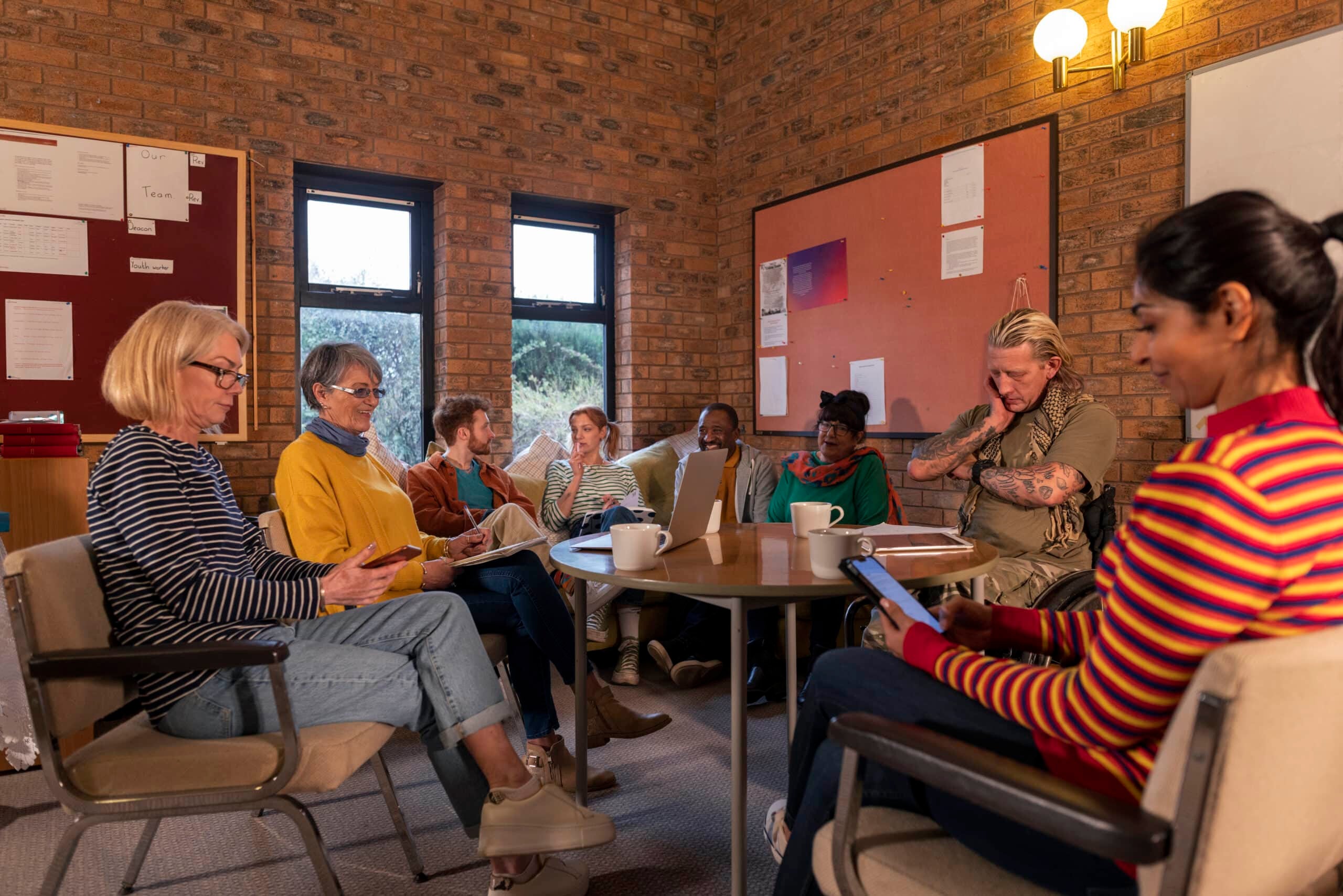 A diverse group of adults gathers in a cozy, brick-walled meeting room, engaged in a discussion with coffee cups, notebooks, and devices on the table.