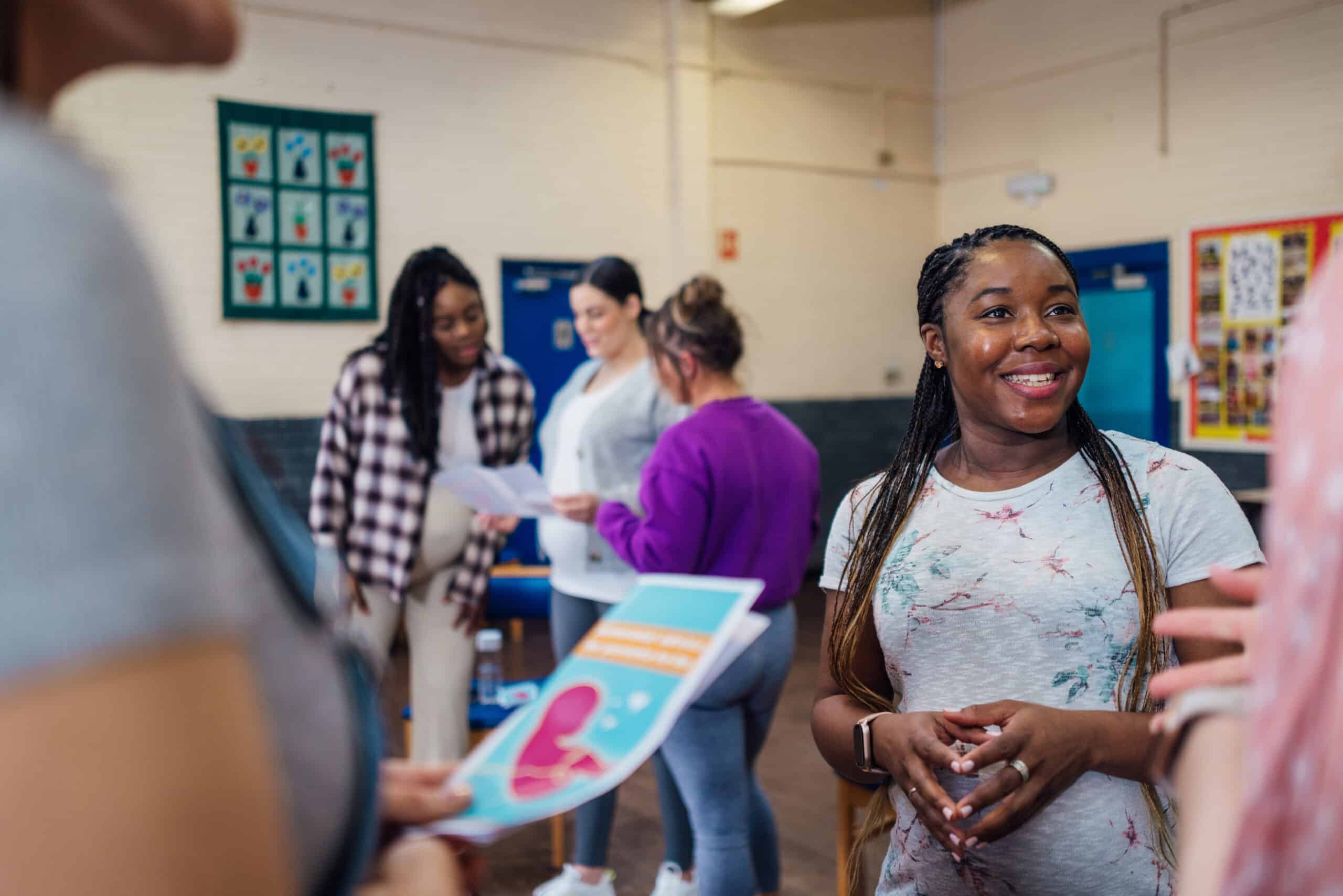 A young woman smiles while participating in a casual group discussion in a classroom setting, with colorful handouts and others conversing in the background.
