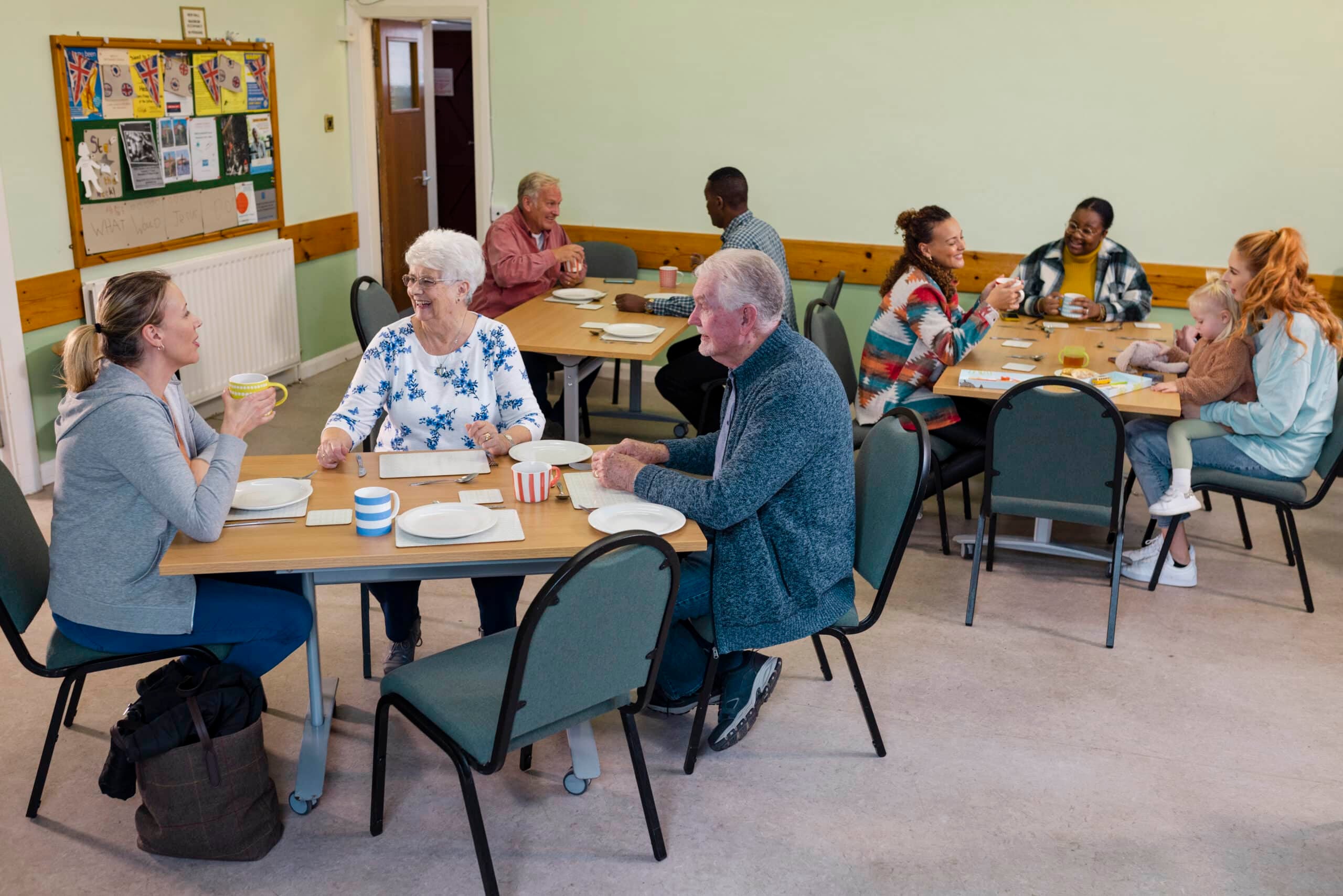 People of different ages gather around tables in a community hall, enjoying drinks and conversation in a relaxed, intergenerational setting.