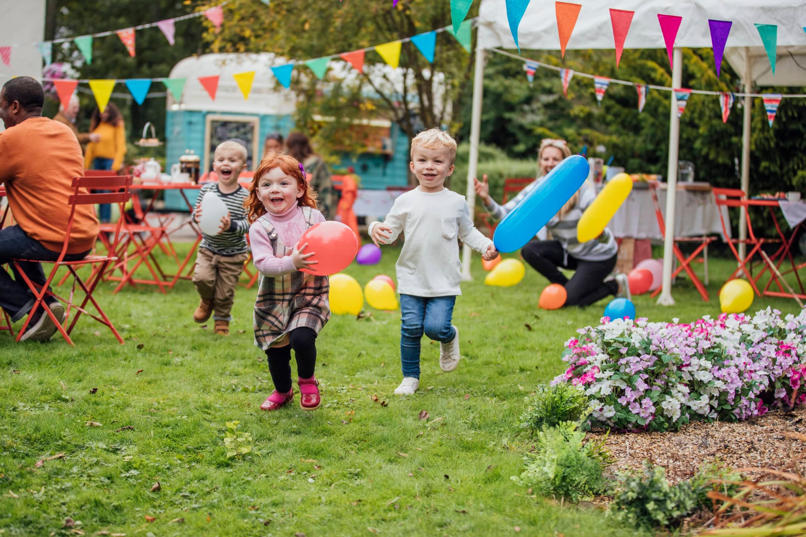 Young children holding colorful balloons run joyfully across a grassy yard during an outdoor festival with bunting, flowers, and picnic tables in the background.