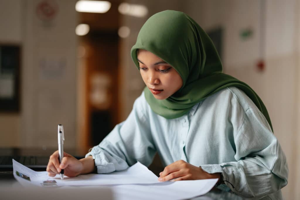 Portrait of a young woman wearing hijab, sitting and writing and assignment for a school project or homework. There is a laptop computer on the desk.