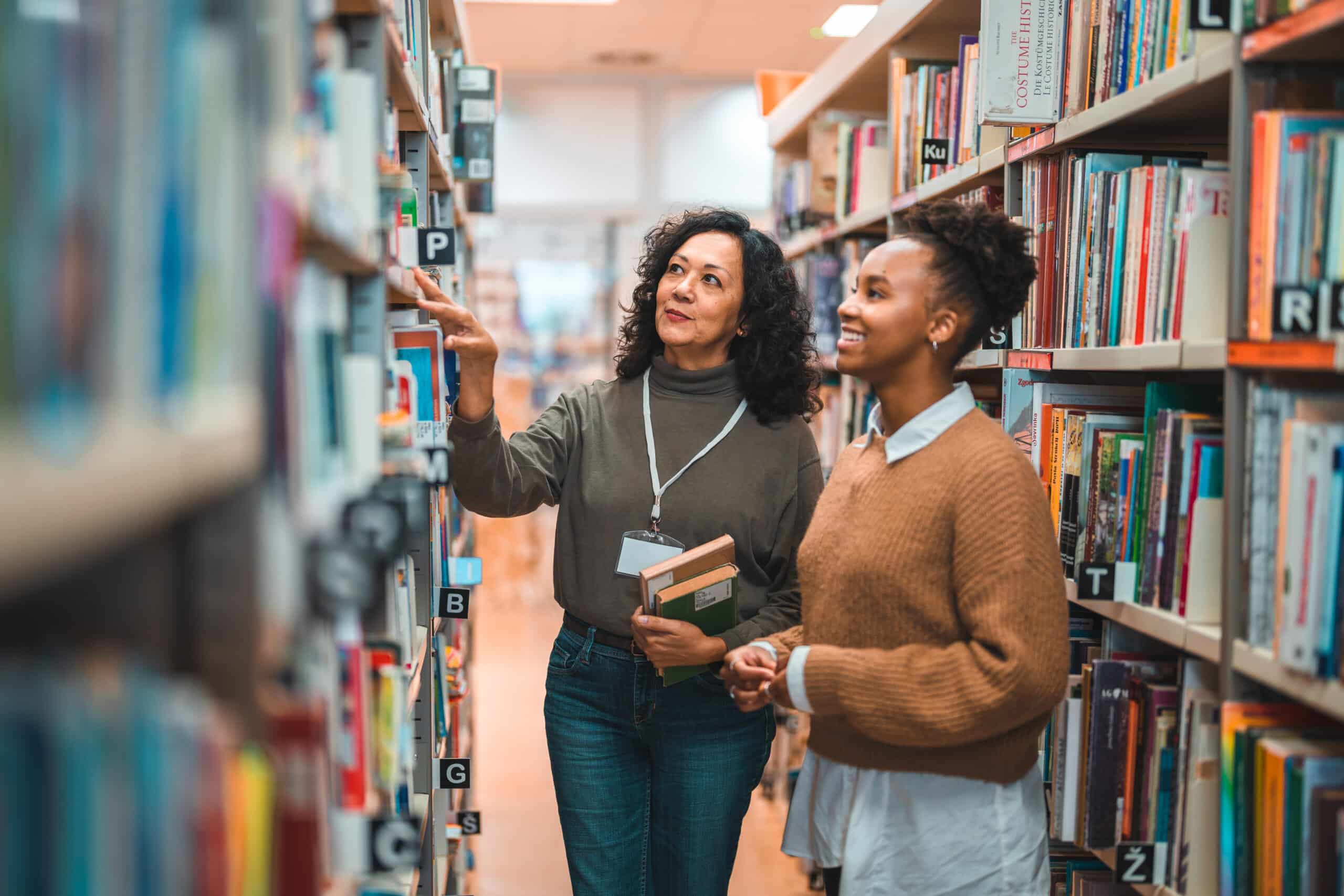 Mid adult Hispanic librarian escorting a student in a library. Searching for a book on shelve.